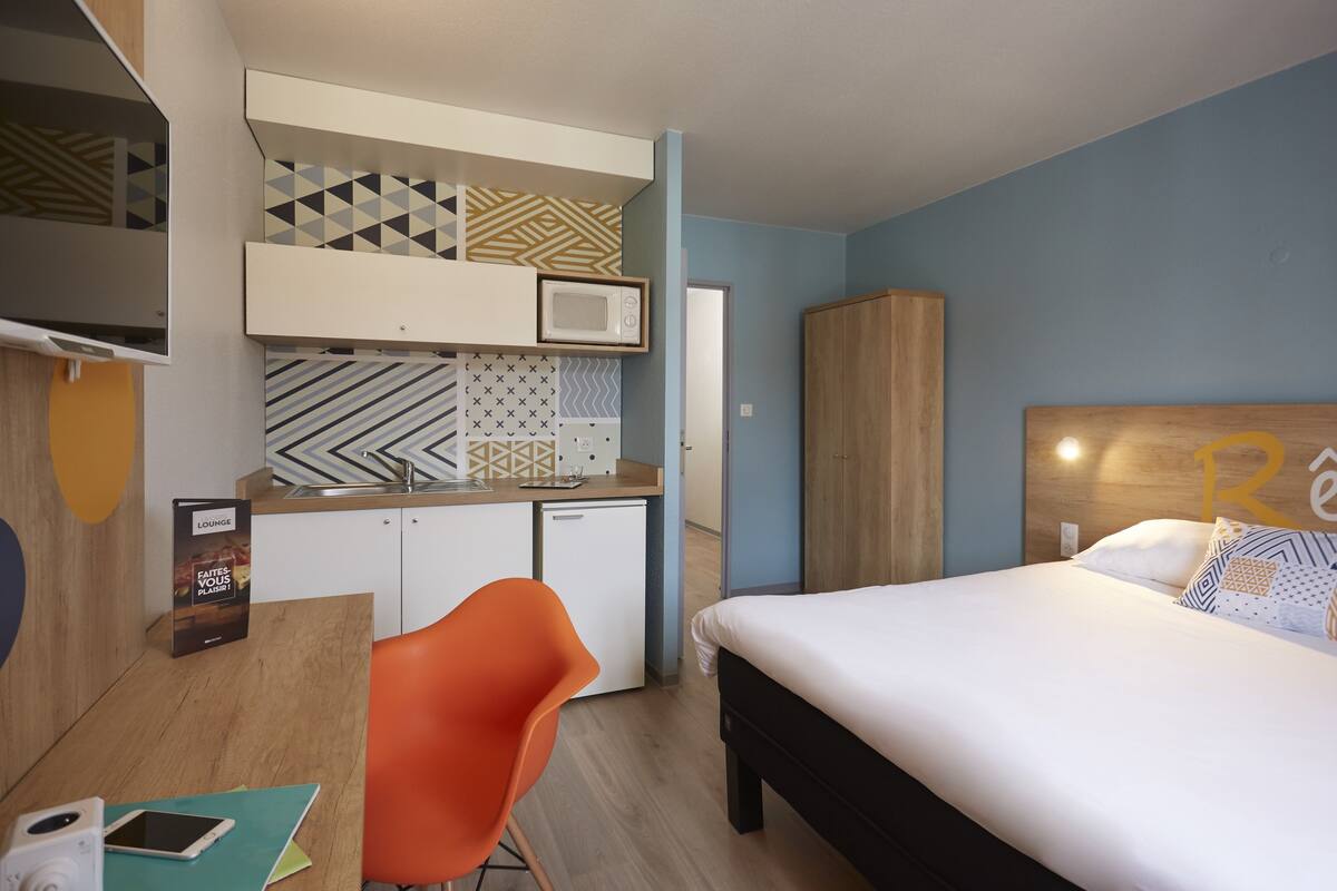 Ein modernes Hotelzimmer mit Bett, Schreibtisch mit Stuhl, Mikrowelle und einer kleinen Küchenzeile.