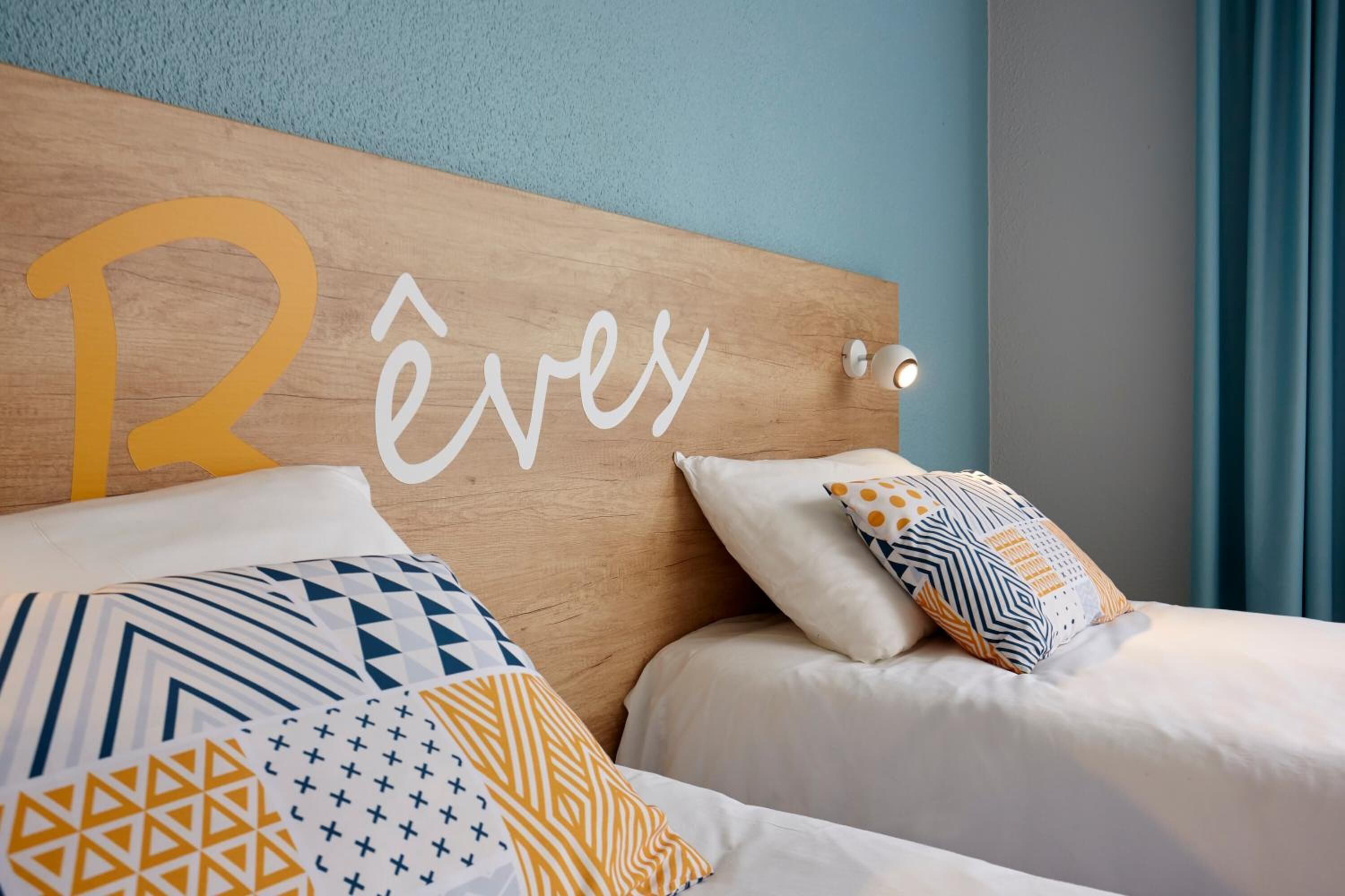 A hotel room with a wooden headboard displaying the name 'Réves'. The room has two beds with patterned bedding and decorative pillows.