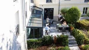 Un área de café al aire libre con mesas y sillas, rodeada de follaje y un edificio con balcones.