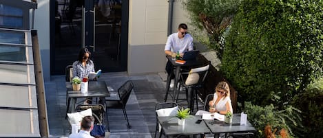 An outdoor seating area with tables and chairs, surrounded by greenery and a building entrance.