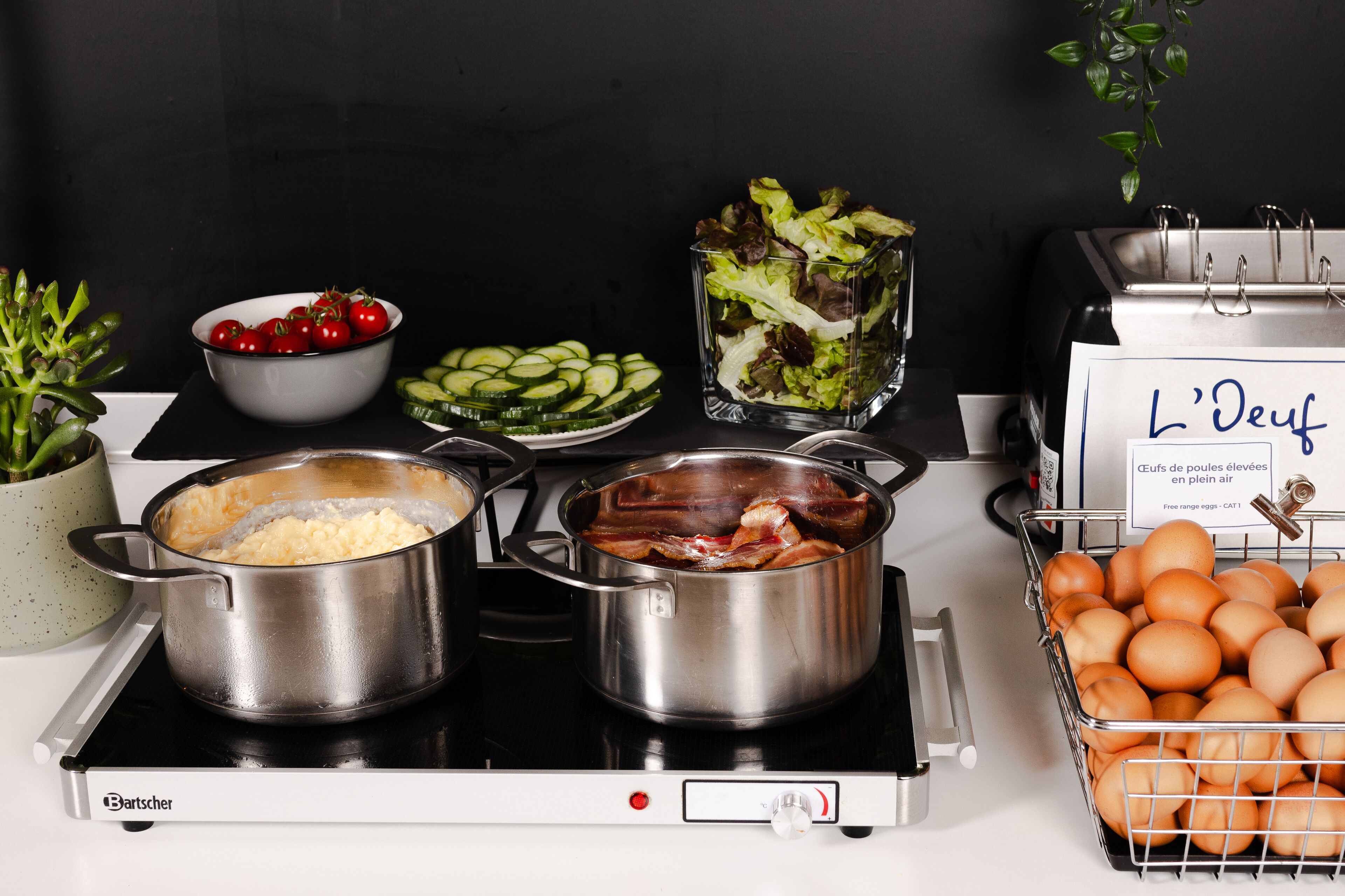 Une cuisine avec deux casseroles sur la cuisinière, un bol de cerises, un bol de concombres coupés en tranches, un bol de laitue et un panier d’œufs.