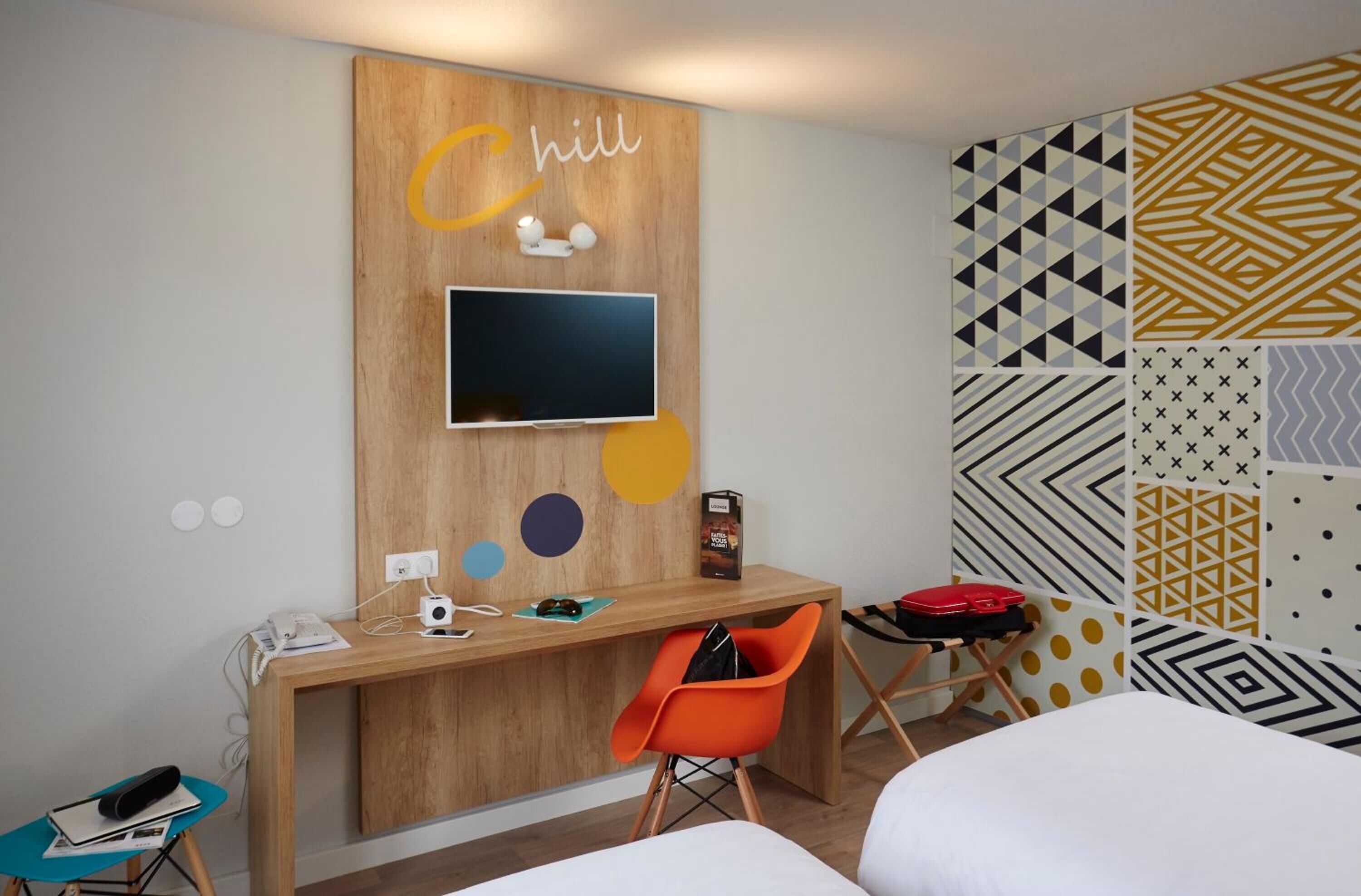 A hotel room with a wooden desk, a flat-screen TV, an orange chair, and a bed.