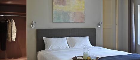 A neatly made bed with white linens, a tray with a glass of water and a lemon, and a painting on the wall.