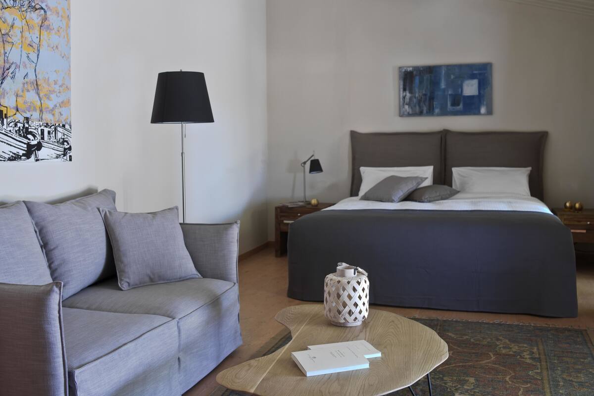 Ein modernes Hotelzimmer mit einem grauen Sofa, einem großen Bett mit grauer Bettwäsche, einem runden Couchtisch mit einem Buch, einer schwarzen Stehlampe und einem Bild an der Wand.