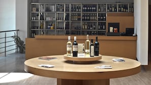 Un espace bar moderne avec une table ronde en bois où sont exposées différentes bouteilles et différents verres. Derrière le bar, on aperçoit un mur où sont rangées de manière ordonnée des bouteilles d’alcool.