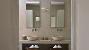 Une salle de bain avec deux lavabos, chacun doté d’un vasque et d’un miroir au-dessus. On y trouve des paniers en osier sous les lavabos et une petite étagère avec des articles de toilette.