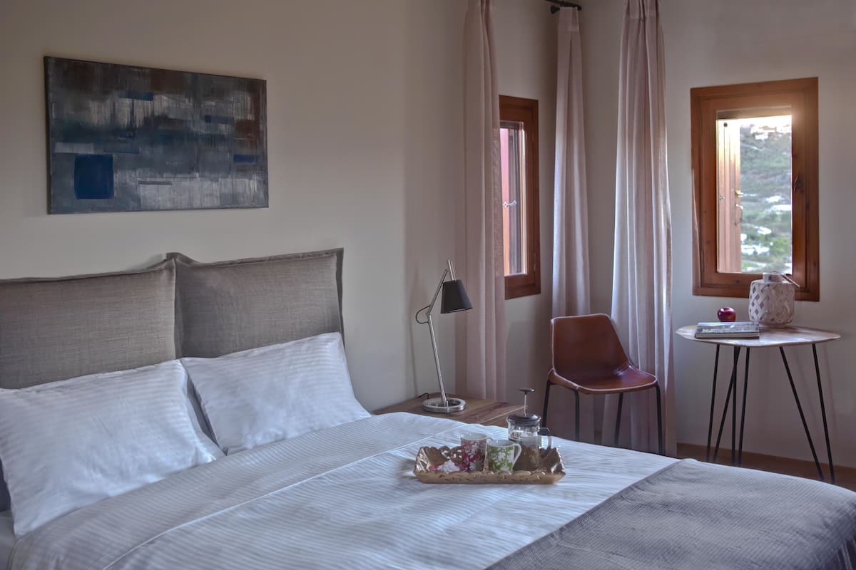 Ein Schlafzimmer mit einem großen Bett, einem kleinen Tisch mit Lampe, einem Stuhl und einem Fenster mit Vorhängen.