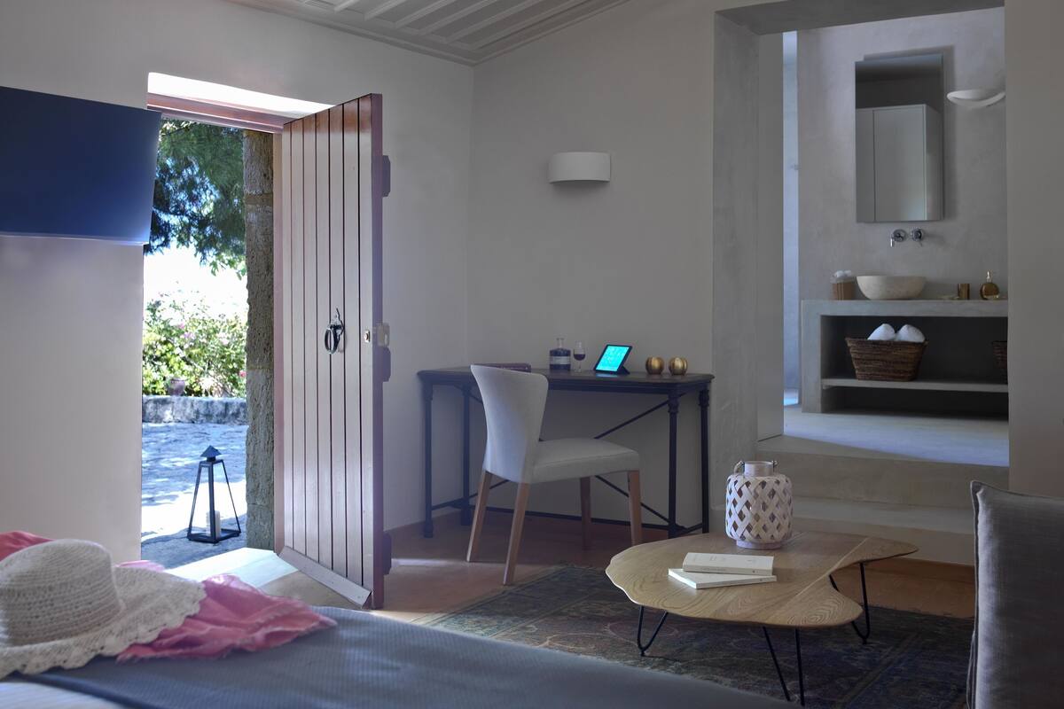Ein Schlafzimmer mit Bett, Schreibtisch mit Laptop, Stuhl, kleinem Tisch mit Buch sowie ein Badezimmer mit Waschbecken und Regalen.