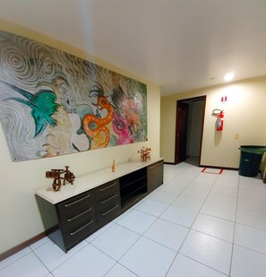 A spacious room with a large abstract painting on the wall, a white tiled floor, a dark-colored cabinet with drawers, and a small toy bicycle on top. - Quaraçá Pousada (Maceió)
