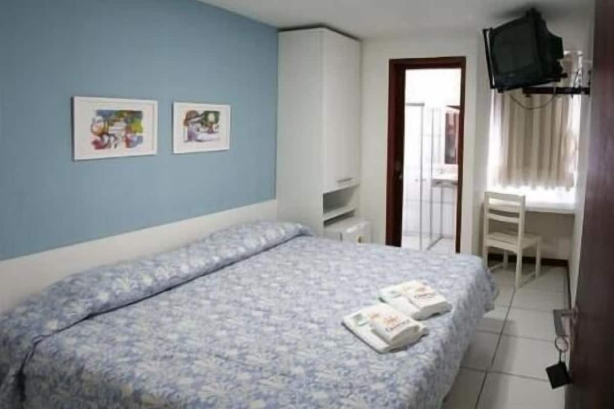 A bedroom with a bed, two framed pictures on the wall, a desk with a chair, and a television mounted on the wall.