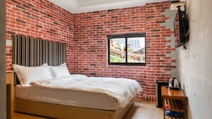 Un dormitorio con pared de ladrillo, una cama, un ventanal con vista a un edificio tradicional y una pequeña mesa de madera con una tetera.