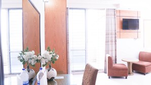 A modern hotel room with a large window, a flat-screen TV mounted on the wall, a sofa, a small table, and decorative vases with flowers. - Hotel Afford Inn (Dhaka)