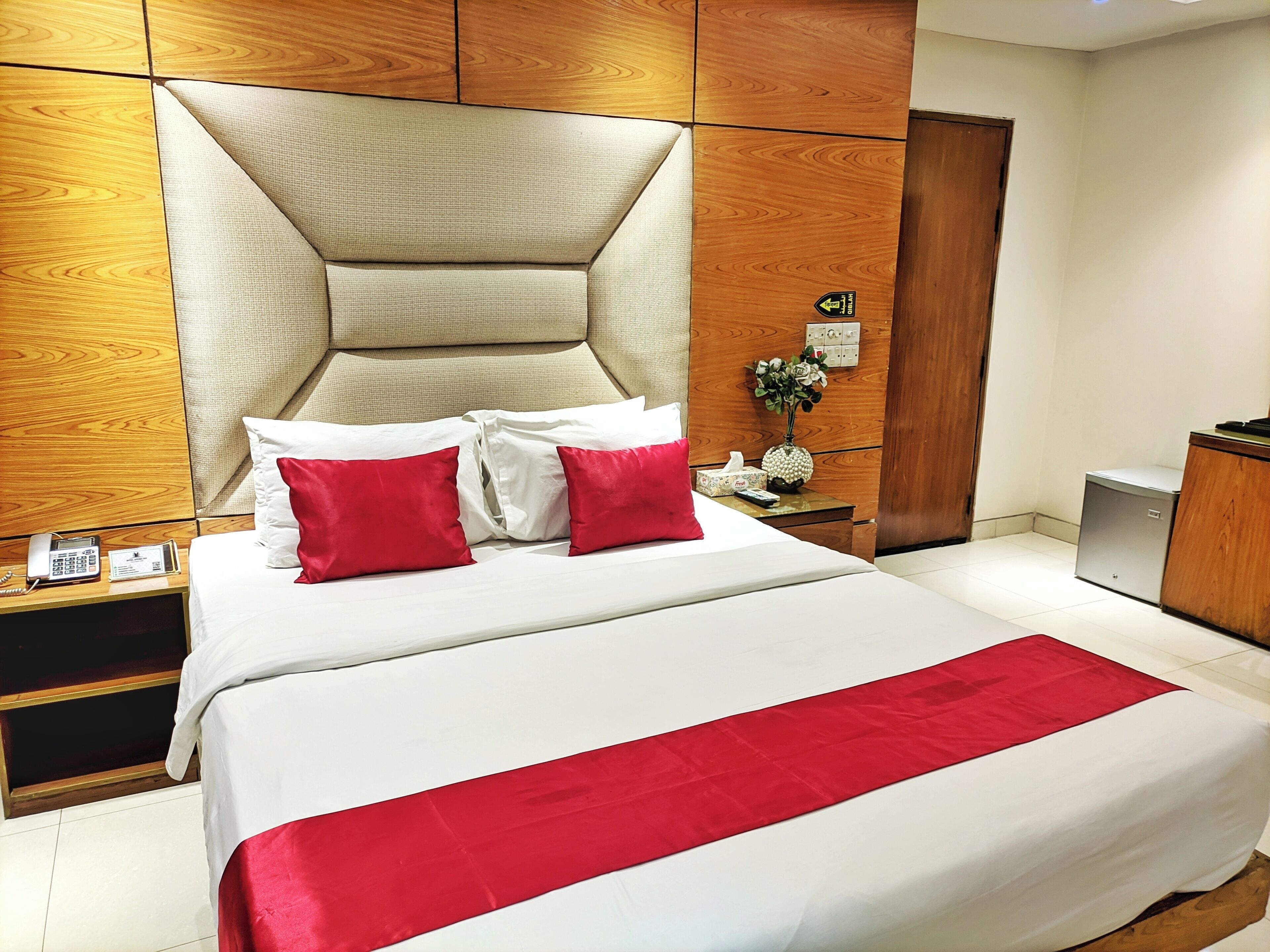 A hotel room with a large bed, a wooden headboard, a bedside table with a phone and tissues, and a small cabinet.