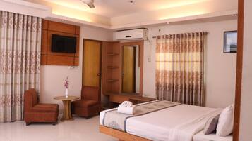 A hotel room with a bed, a TV mounted on the wall, a wardrobe, a small table with a vase, and a chair.