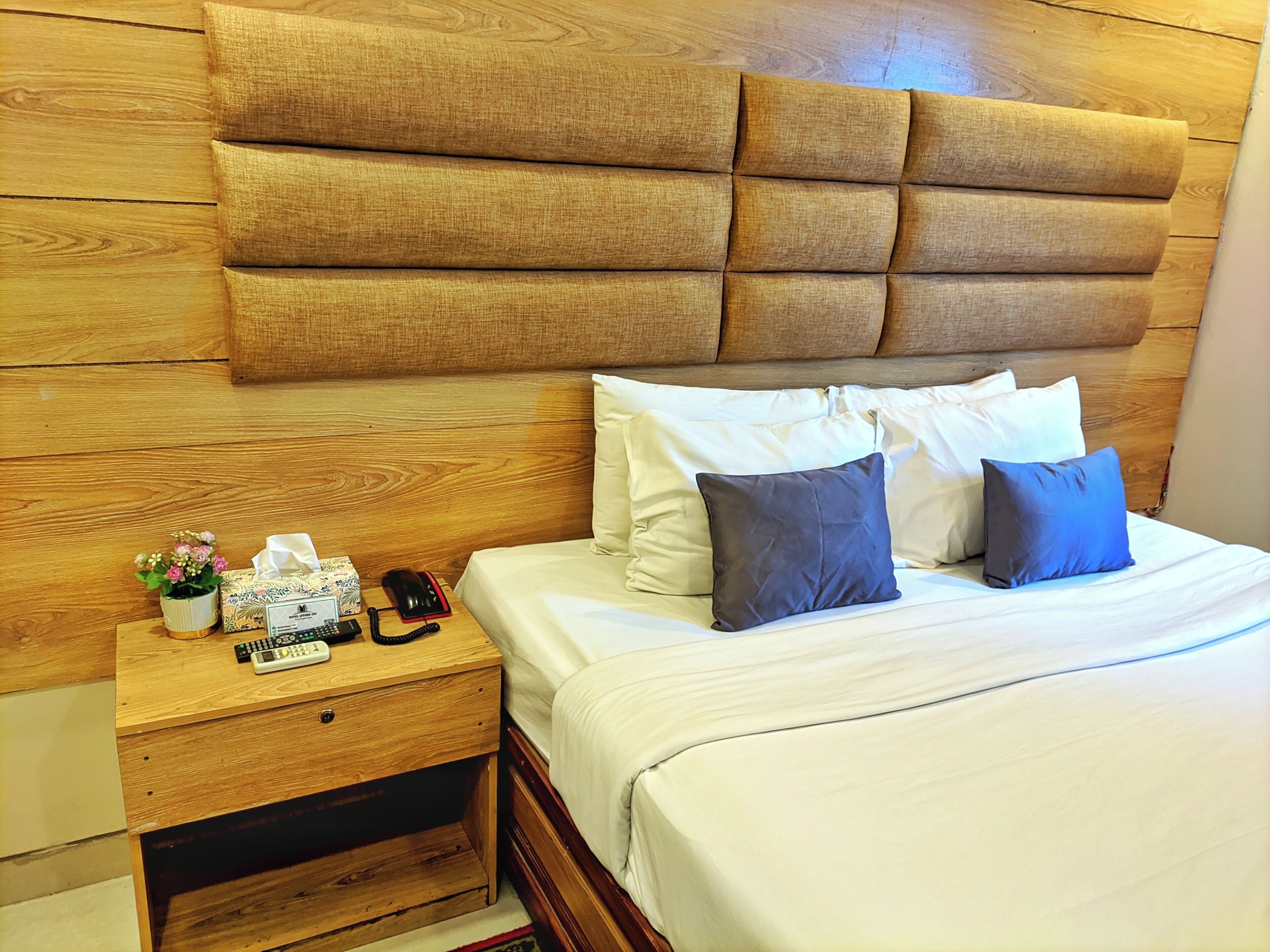 Habitación de hotel con cabecera de madera, mesita de noche con teléfono y controles remotos, y cama con varias almohadas.