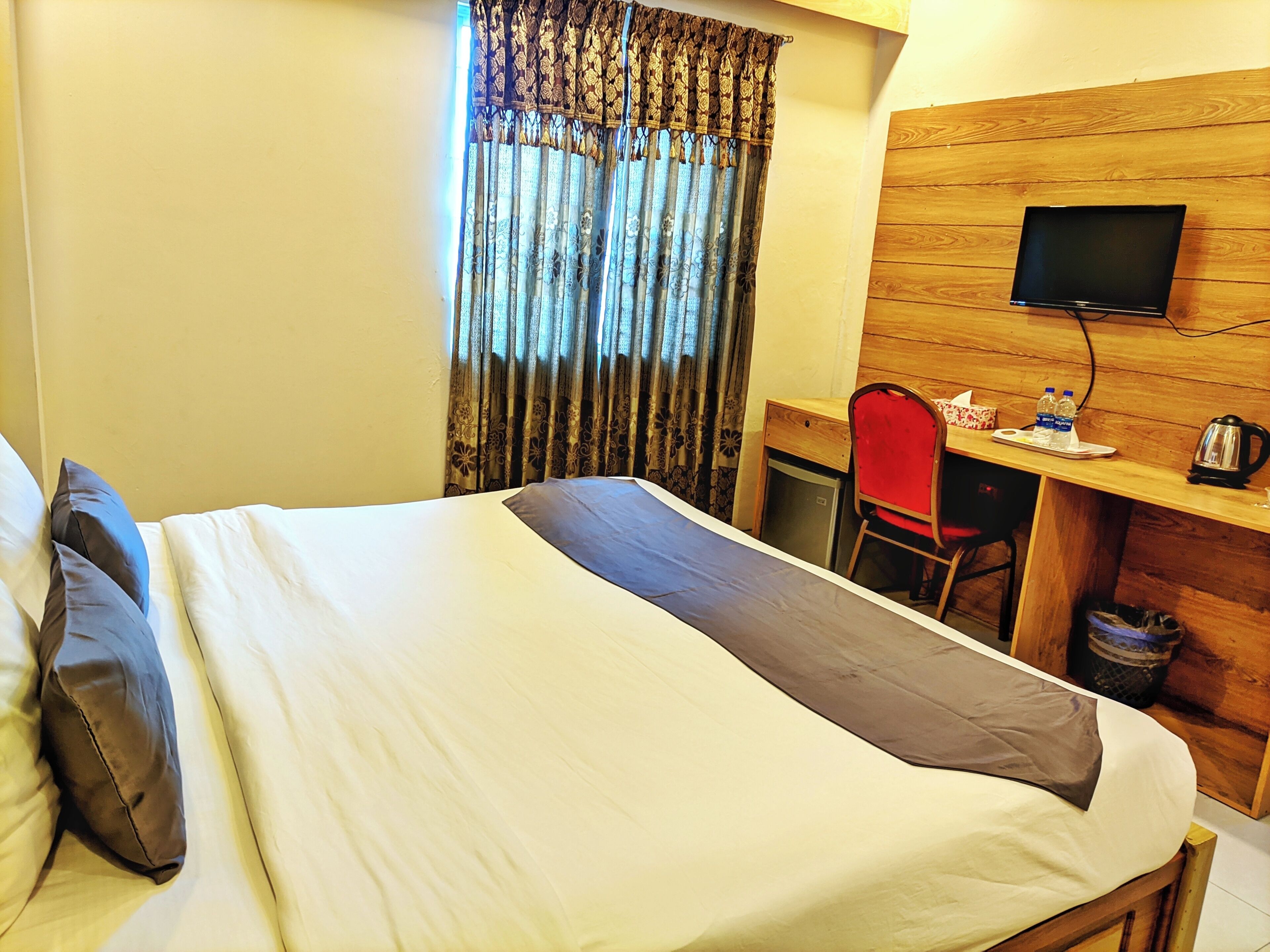 A hotel room with a bed, a desk with a chair, a television, and a small kitchenette area.
