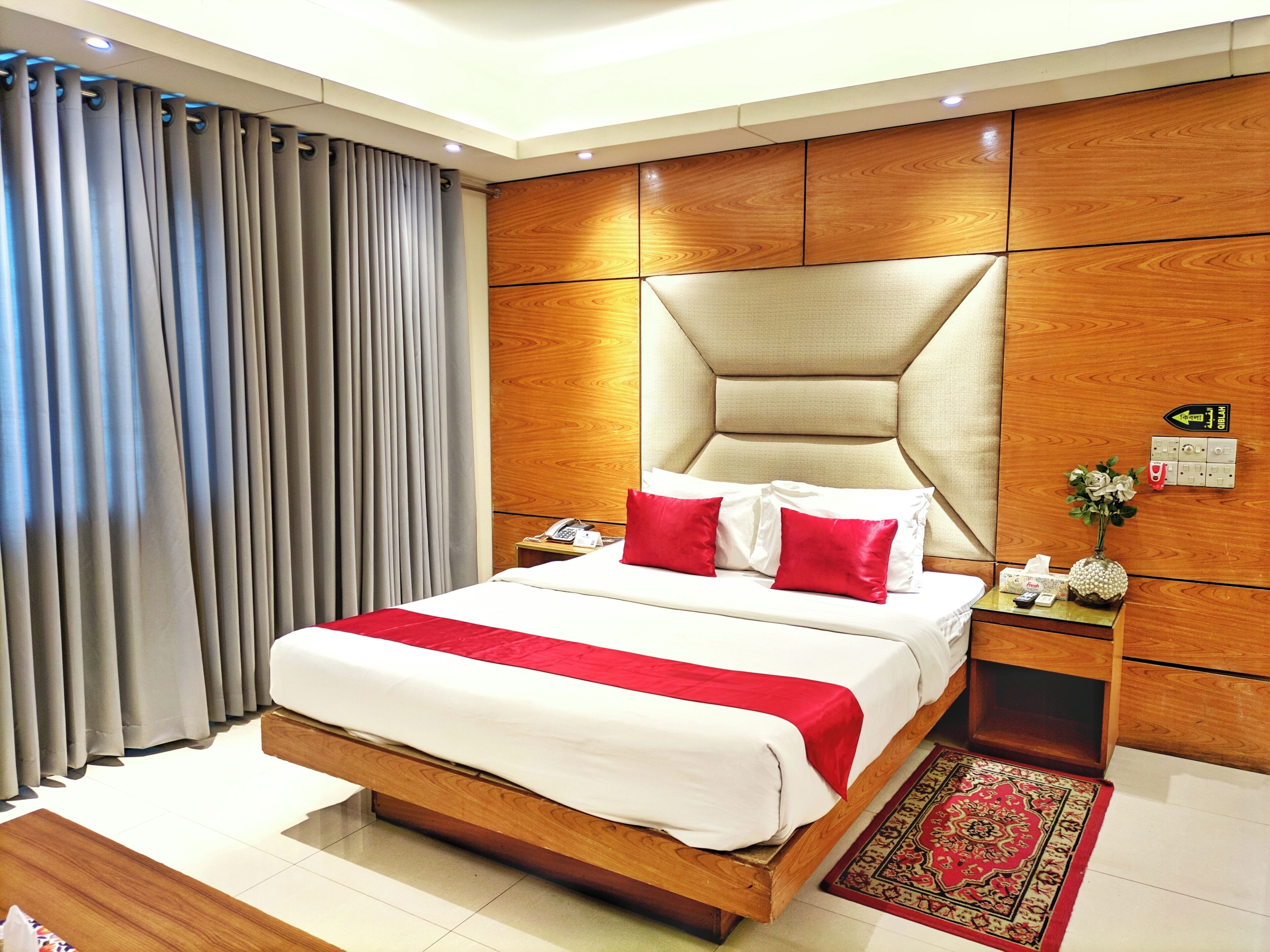 Une chambre d’hôtel avec un grand lit, une tête de lit en bois, des tables de chevet, un téléphone, un vase avec des fleurs et une couvre-lit rouge et blanche.