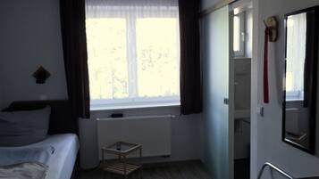 A room with a bed, a small table, a chair, a radiator, a window with curtains, and a mirror on the wall.