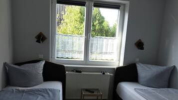 A small, well-lit room with two beds, a radiator, a small table, and a window with a view of trees.