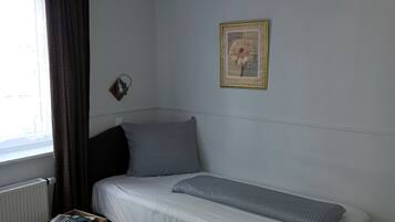Een enkel bed met hoofdeinde, een klein houten nachtkastje met afstandsbediening en een boek, een lijst met een afbeelding aan de muur, en een raam met gordijnen.