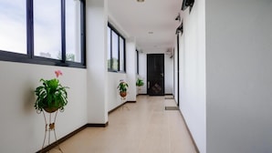 Een lange, smalle gang met grote ramen aan één kant, waardoor natuurlijk licht binnenvalt. Er staan potplanten langs de gang, en een deur aan het einde.