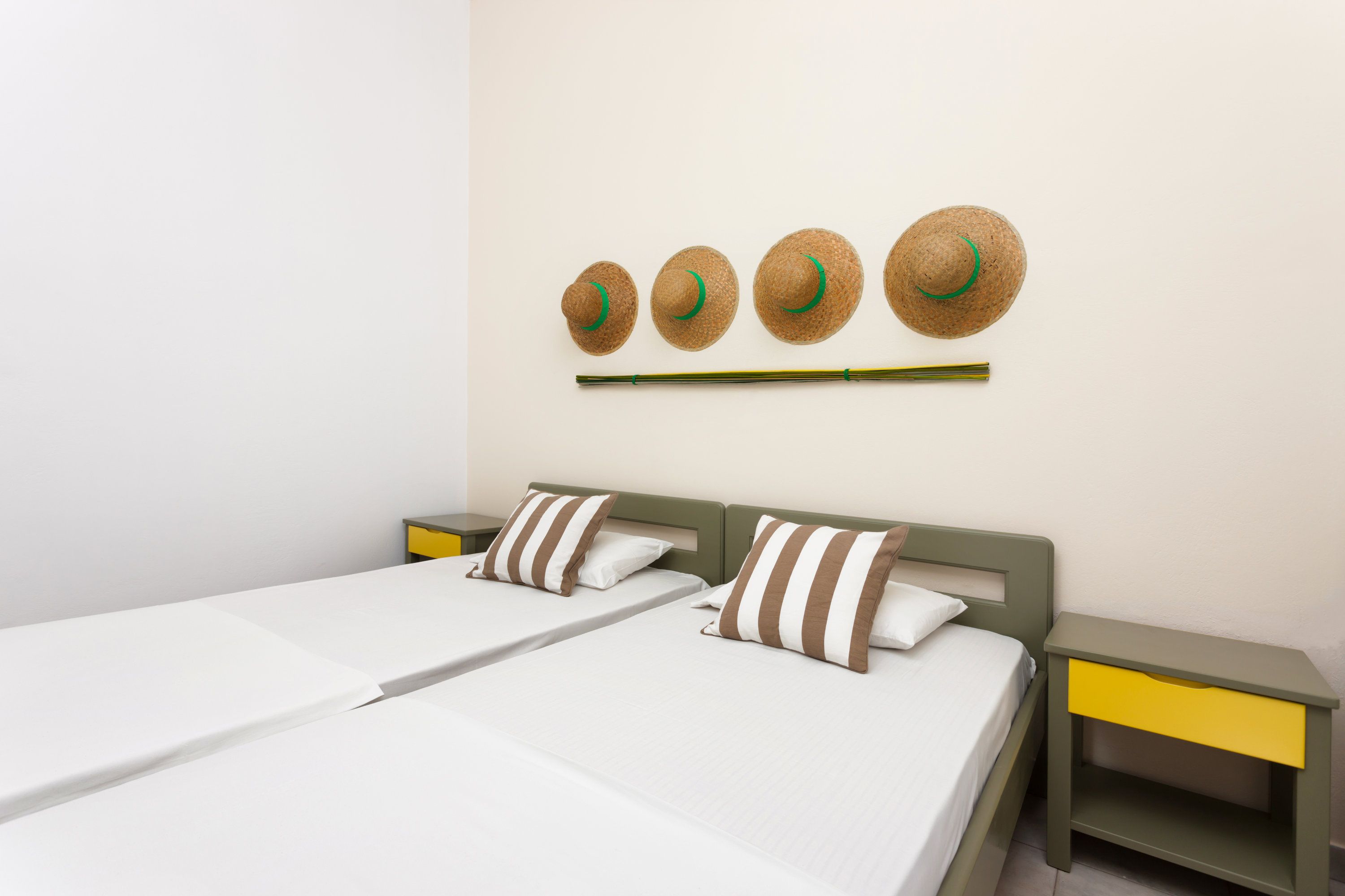 Habitación de hotel con dos camas, una mesita de noche con cajón amarillo y un estante en la pared con cuatro sombreros.