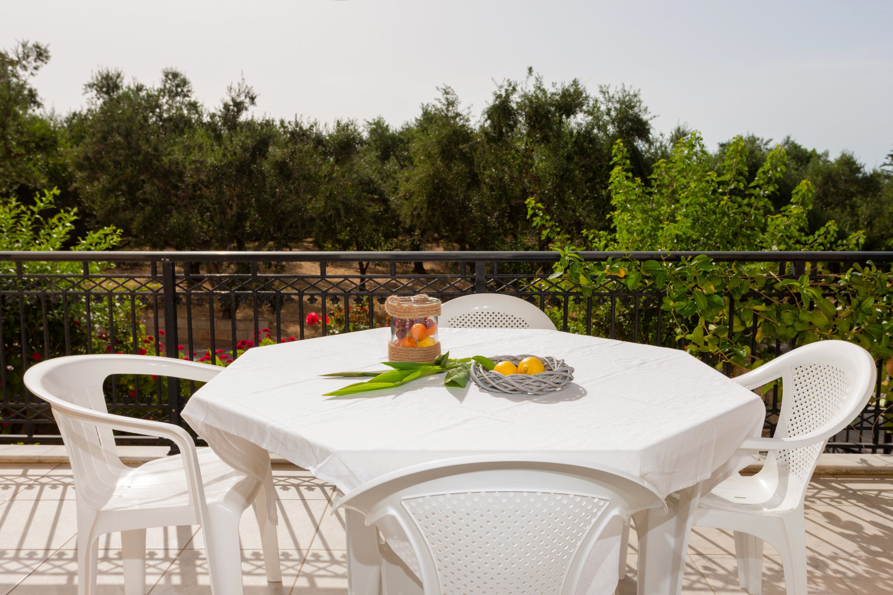 A round table with a white tablecloth, surrounded by white chairs, set on a balcony with a metal railing and greenery in the background.