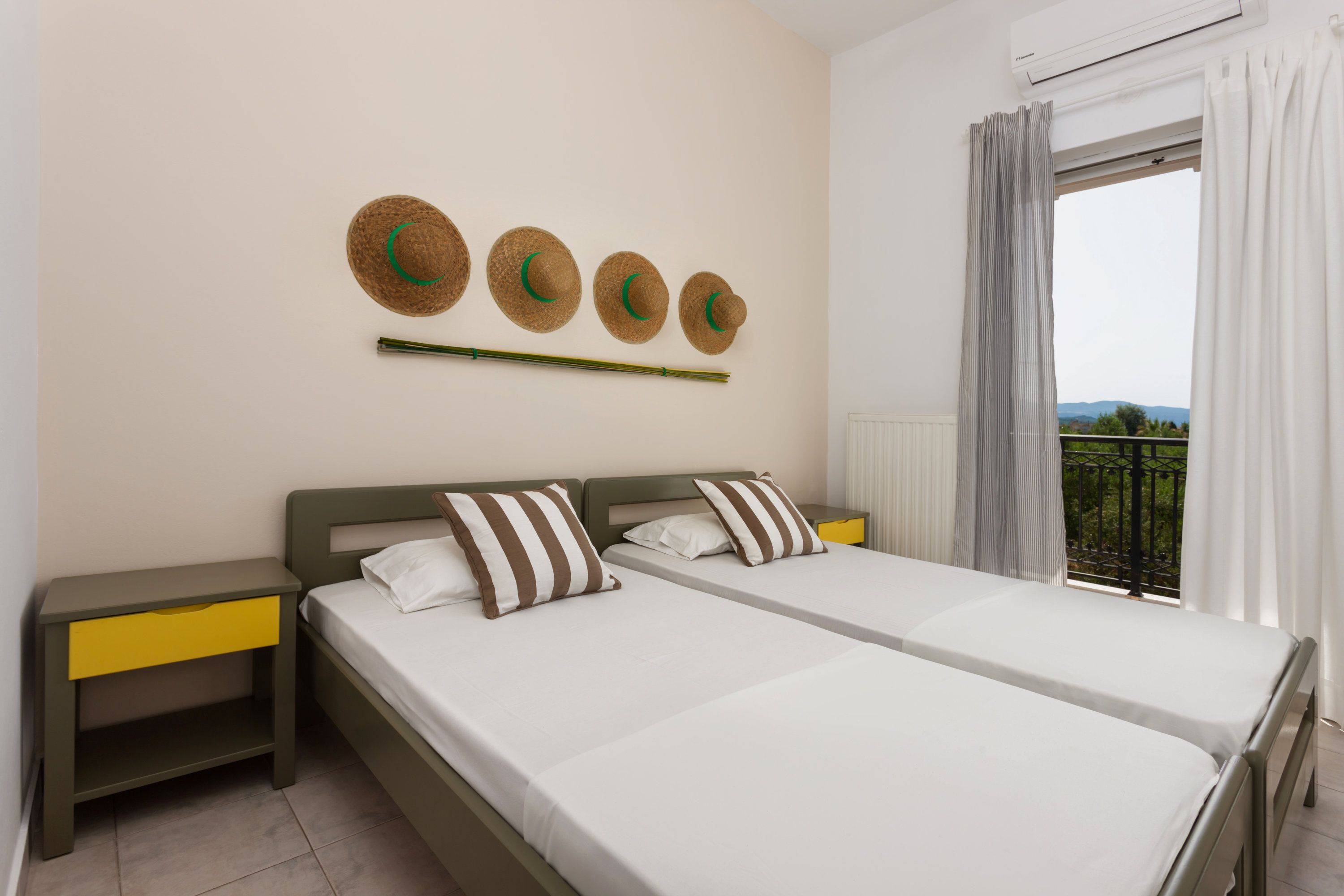 A double bed with white bedding and striped pillows, a nightstand with a yellow drawer, and a wall-mounted hat display.