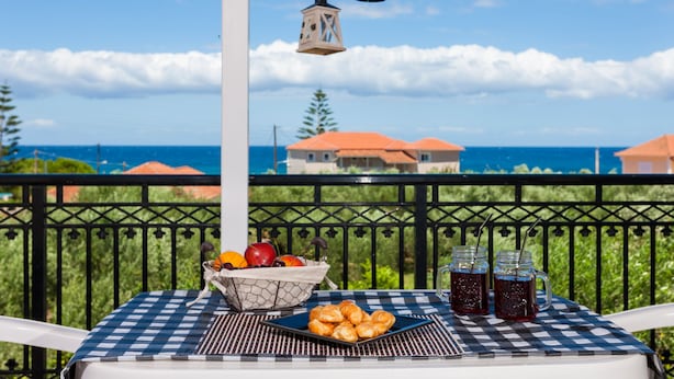 A balcony with a checkered tablecloth, a basket of fruits, a plate of pastries, and two mason jars of beverages. - Galatea (Zakynthos)