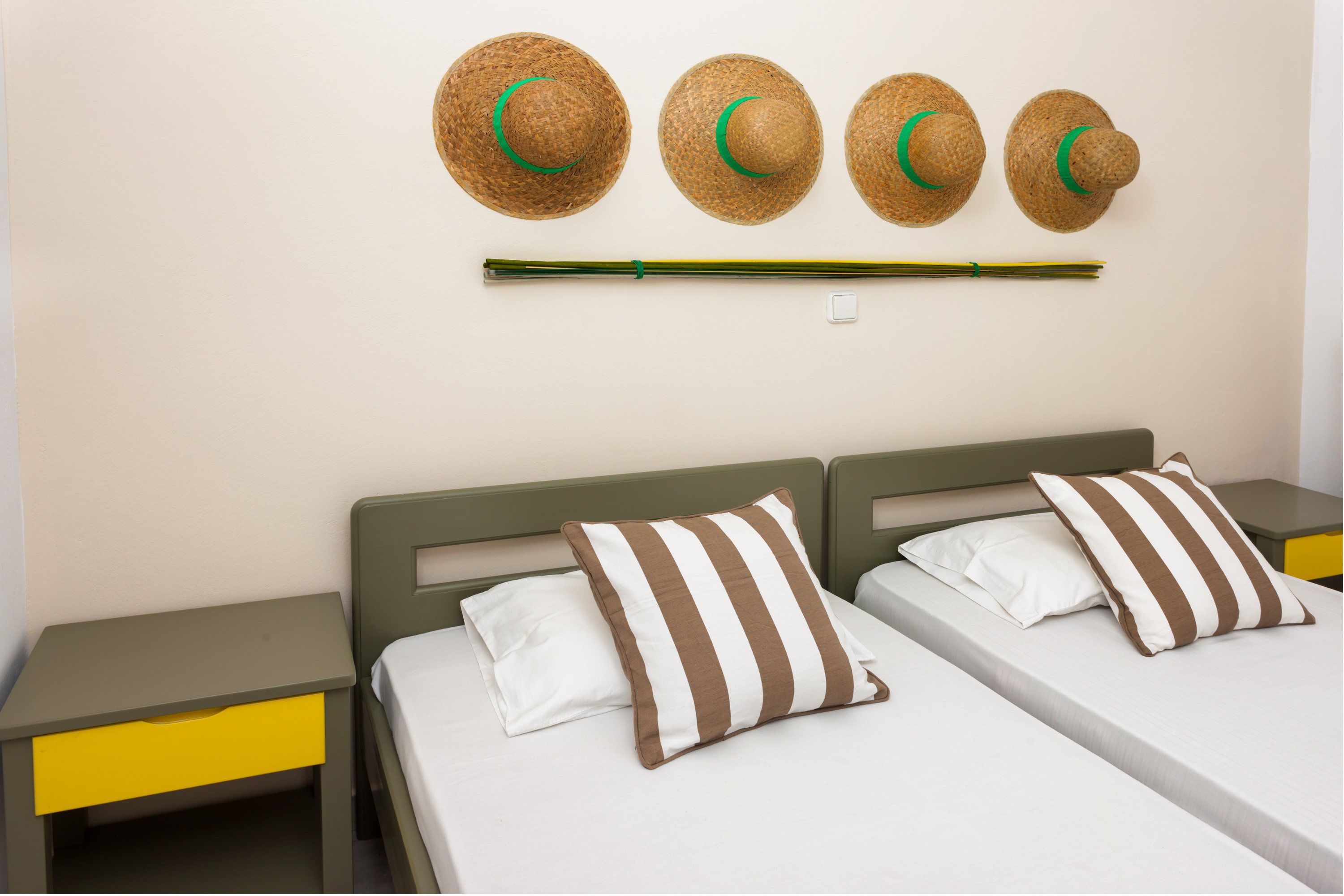 Quarto de hotel com duas camas, cada uma com almofadas listradas, uma mesa de cabeceira com gaveta e um porta-chapéus fixado na parede, com chapéus.
