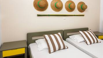 Quarto de hotel com duas camas, cada uma com almofadas listradas, uma mesa de cabeceira com gaveta e um porta-chapéus fixado na parede, com chapéus.
