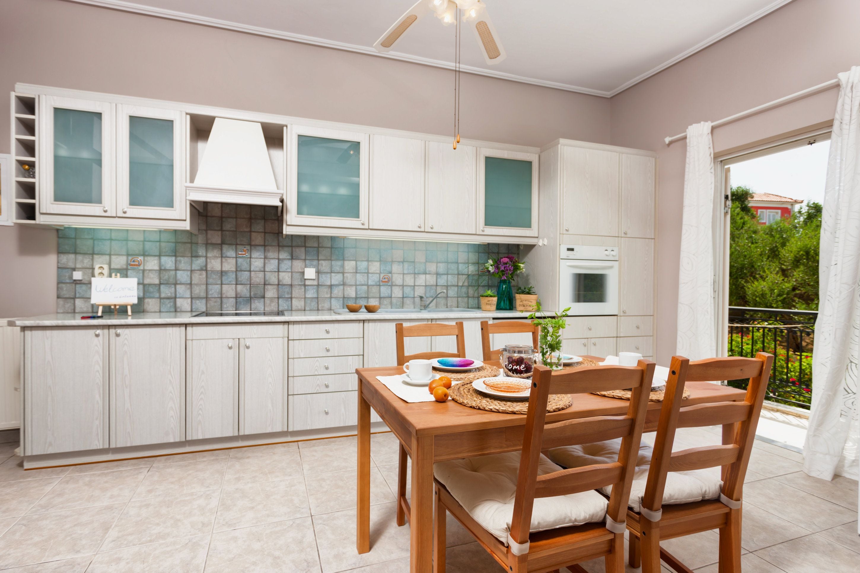 A modern kitchen with white cabinets, a dining table with chairs, and a view of a garden through the window.