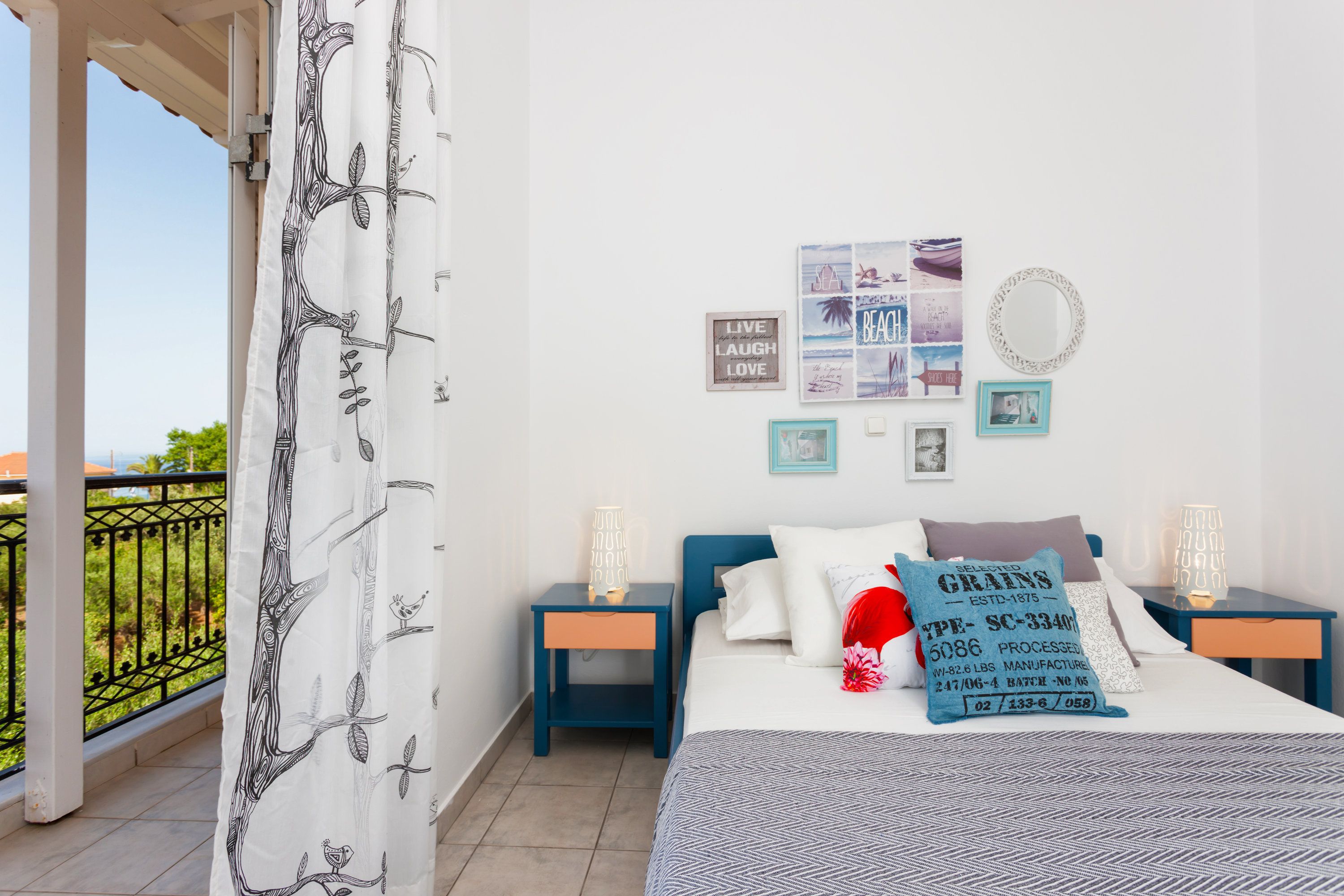 Une chambre avec un lit, deux tables de chevet, un mur orné de tableaux encadrés, un rideau à motifs d’arbres et un balcon avec une balustrade.