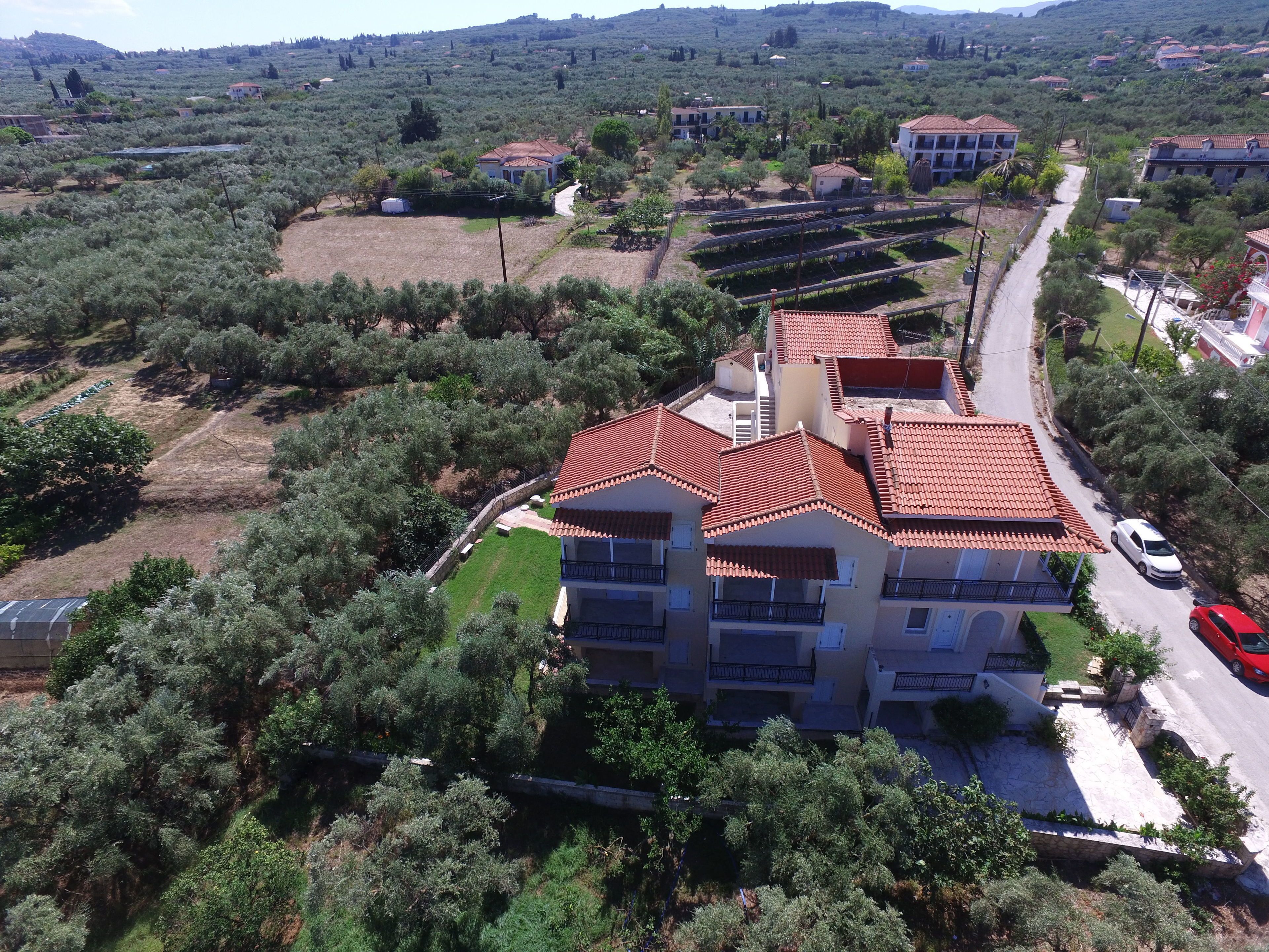 A multi-story building with a red-tiled roof, surrounded by olive trees and a road.