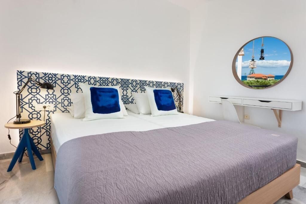Un dormitorio ordenado con un cabecero estampado, una cama con cubrecama gris y un espejo redondo con vistas al mar.
