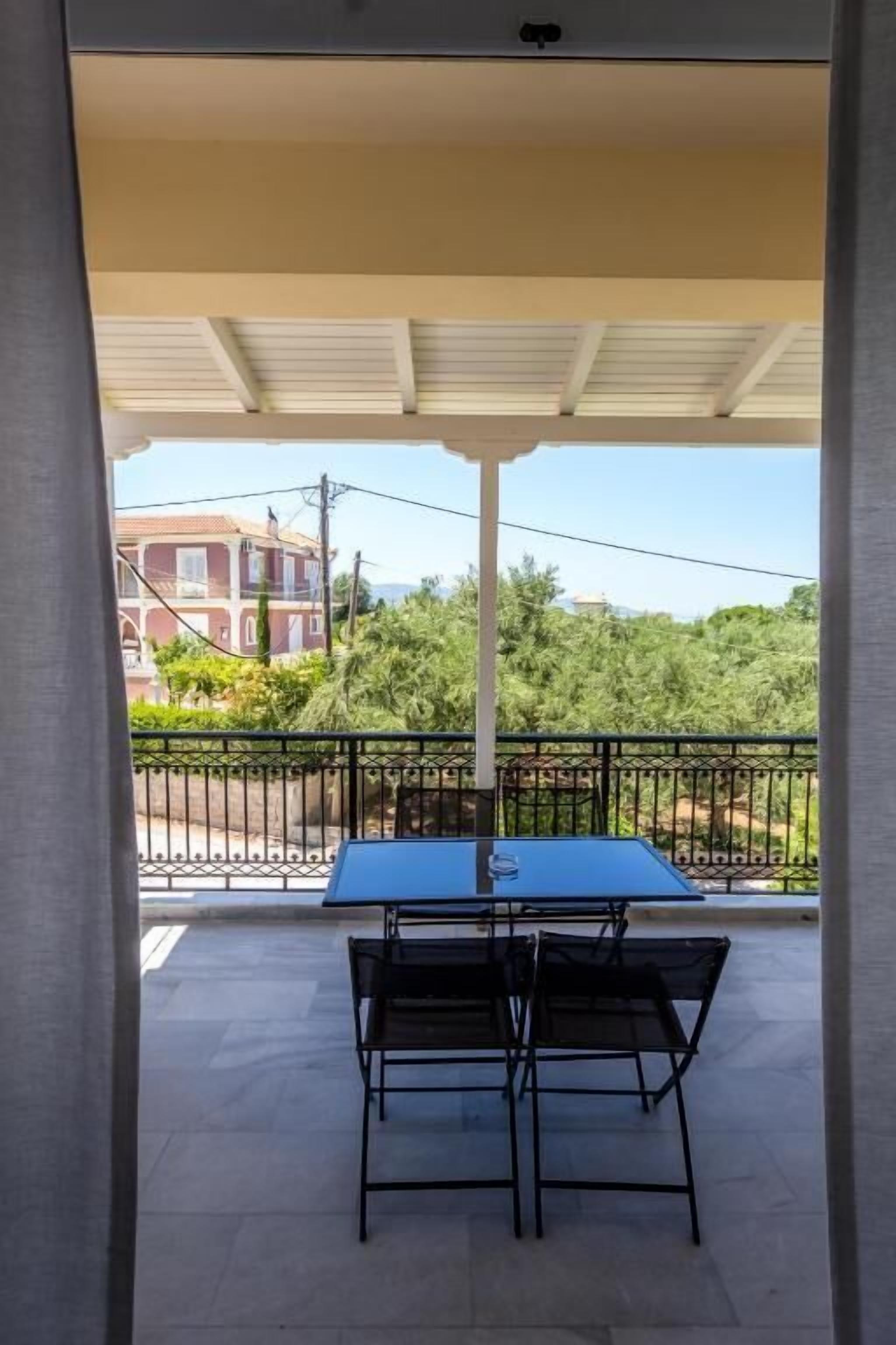 A covered outdoor area with a table and chairs, a view of a building and greenery in the background.