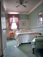 Un dormitorio con cama, una silla, mesita de noche, ventilador de techo, ventana con cortinas y un cuadro en la pared.
