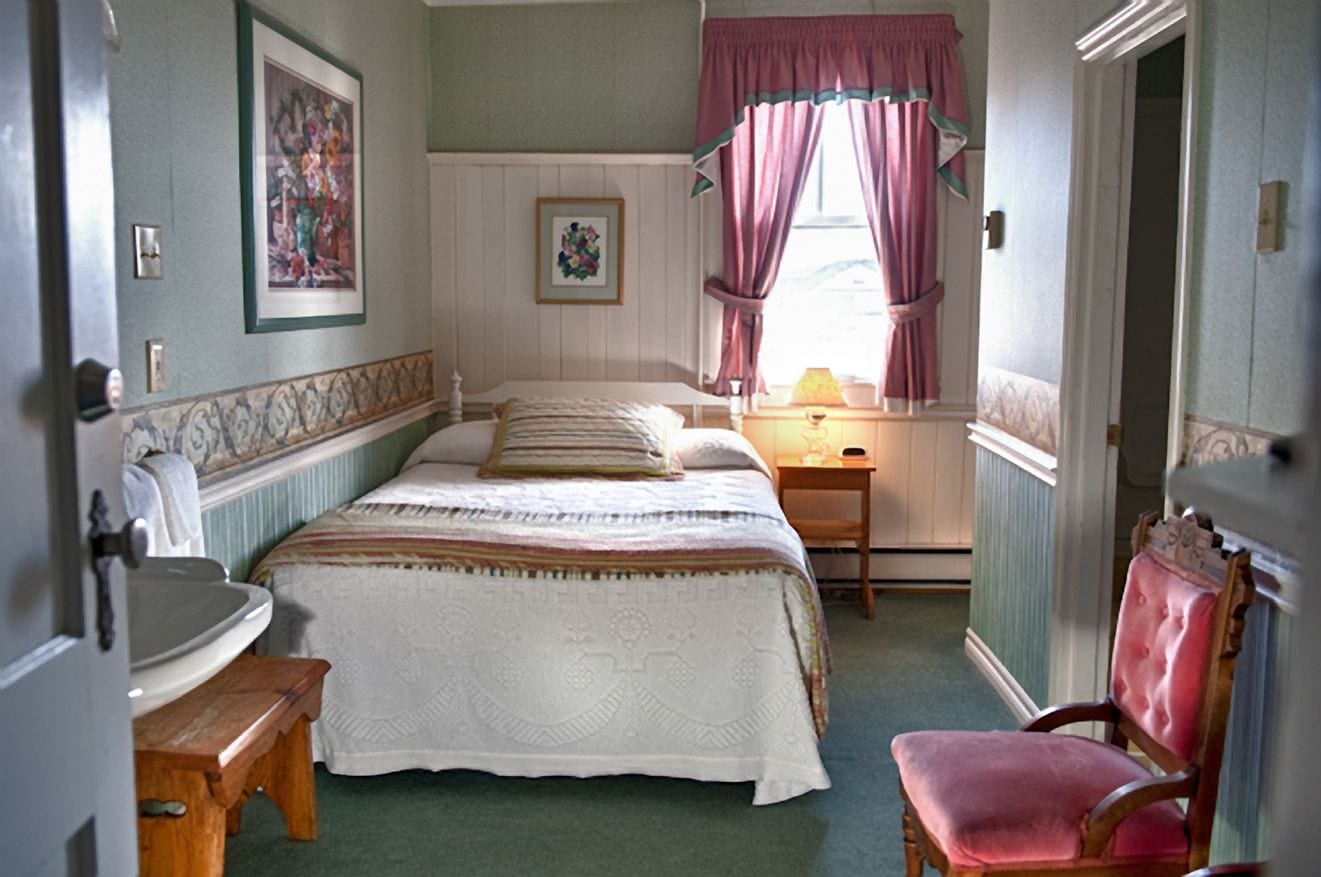 Un dormitorio con cama, mesita de noche, una silla, ventana con cortinas y puerta.