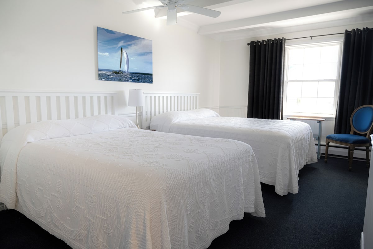 Hotellihuone, jossa on kaksi sänkyä, kattotuuletin, verhoilla varustettu ikkuna ja tuoli.