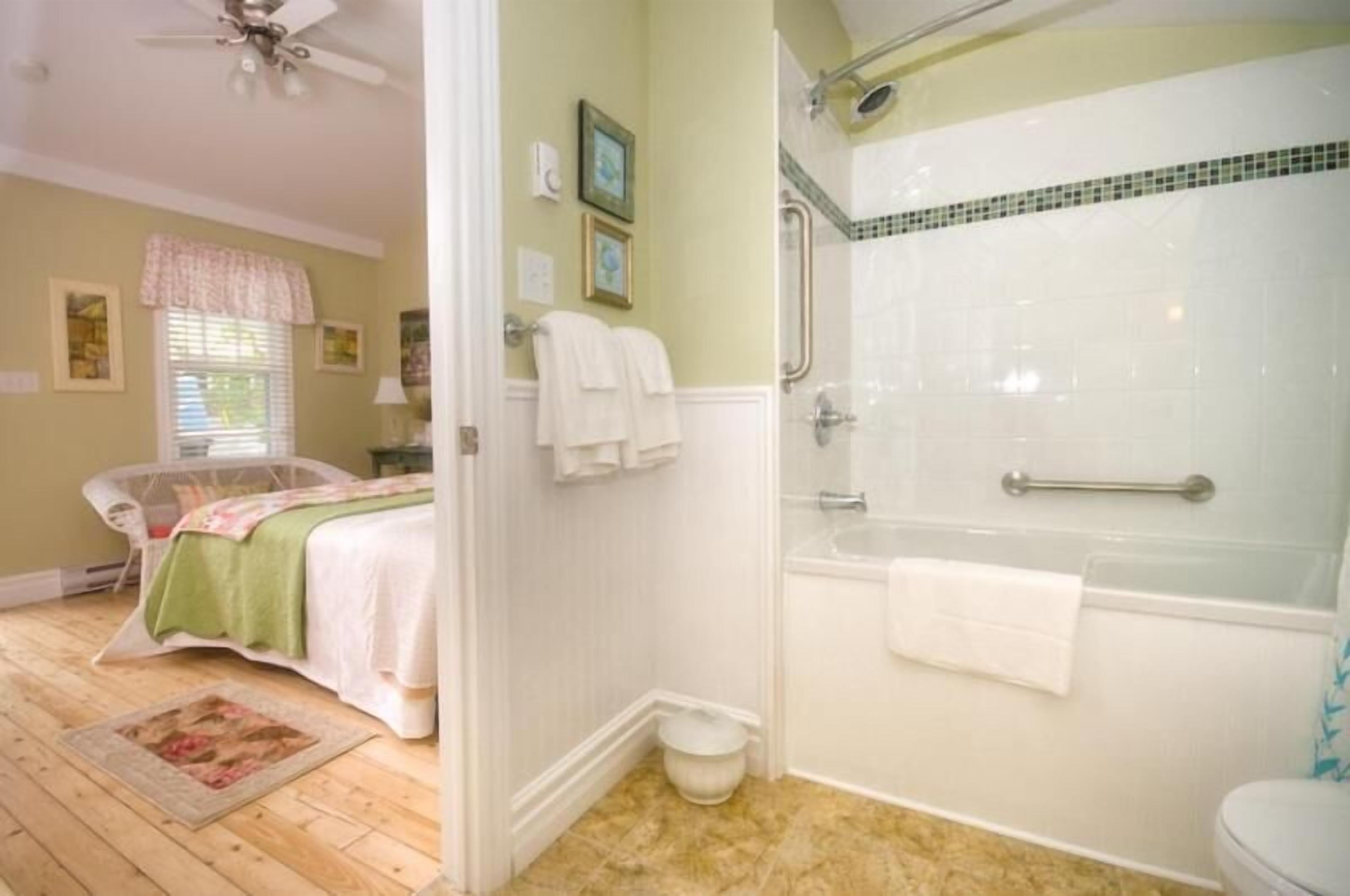 A bathroom with a shower, a toilet, and a sink. There is a window with blinds and a bed with a green blanket.