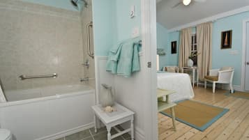 Un baño con bañera, ducha e inodoro. Hay una silla blanca y una mesita con una toalla. Las paredes son azul claro y el suelo es de madera.