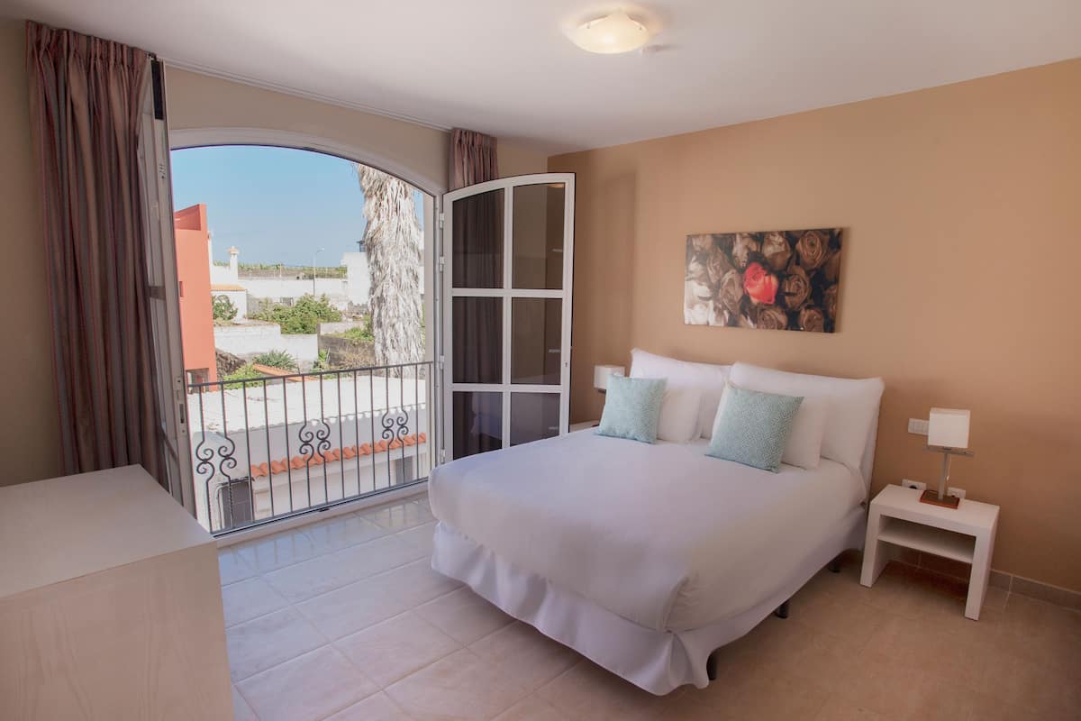Un dormitorio con una cama grande, una mesita de noche, un cuadro y un balcón con vistas a edificios y árboles.