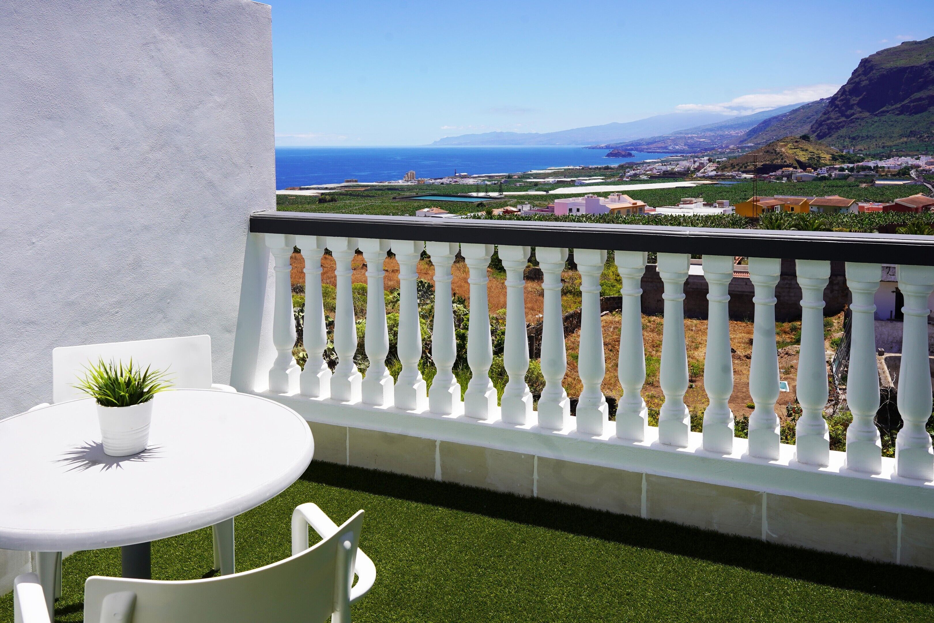 A balcony with a white railing, a small round table with a potted plant, and a view of the sea and distant buildings.
