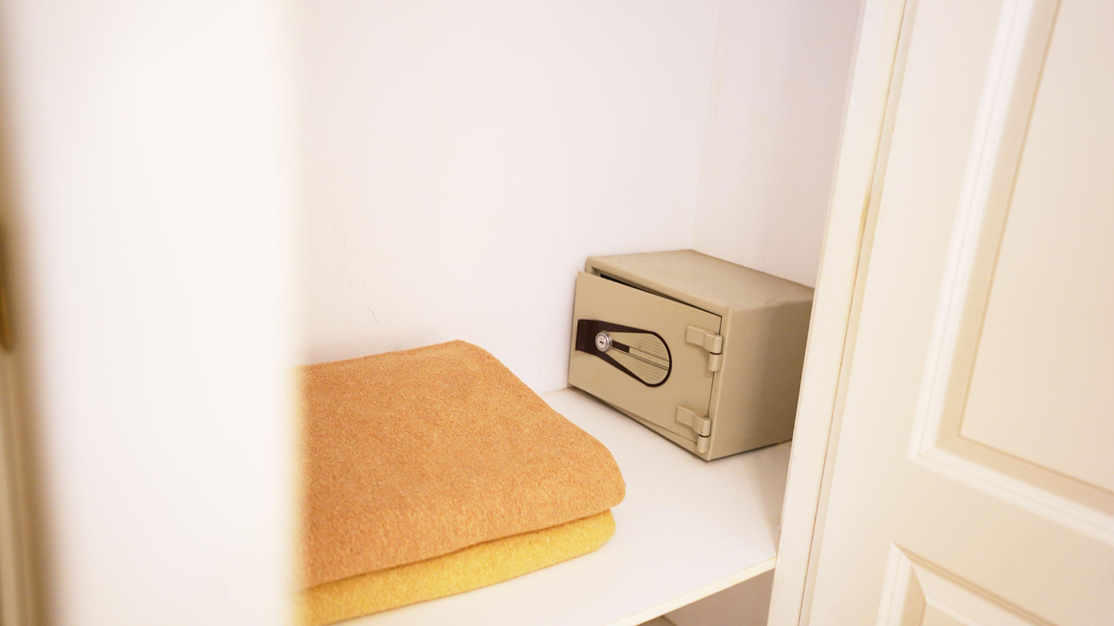 Una piccola cassaforte beige su uno scaffale bianco, un asciugamano beige piegato e una porta bianca.