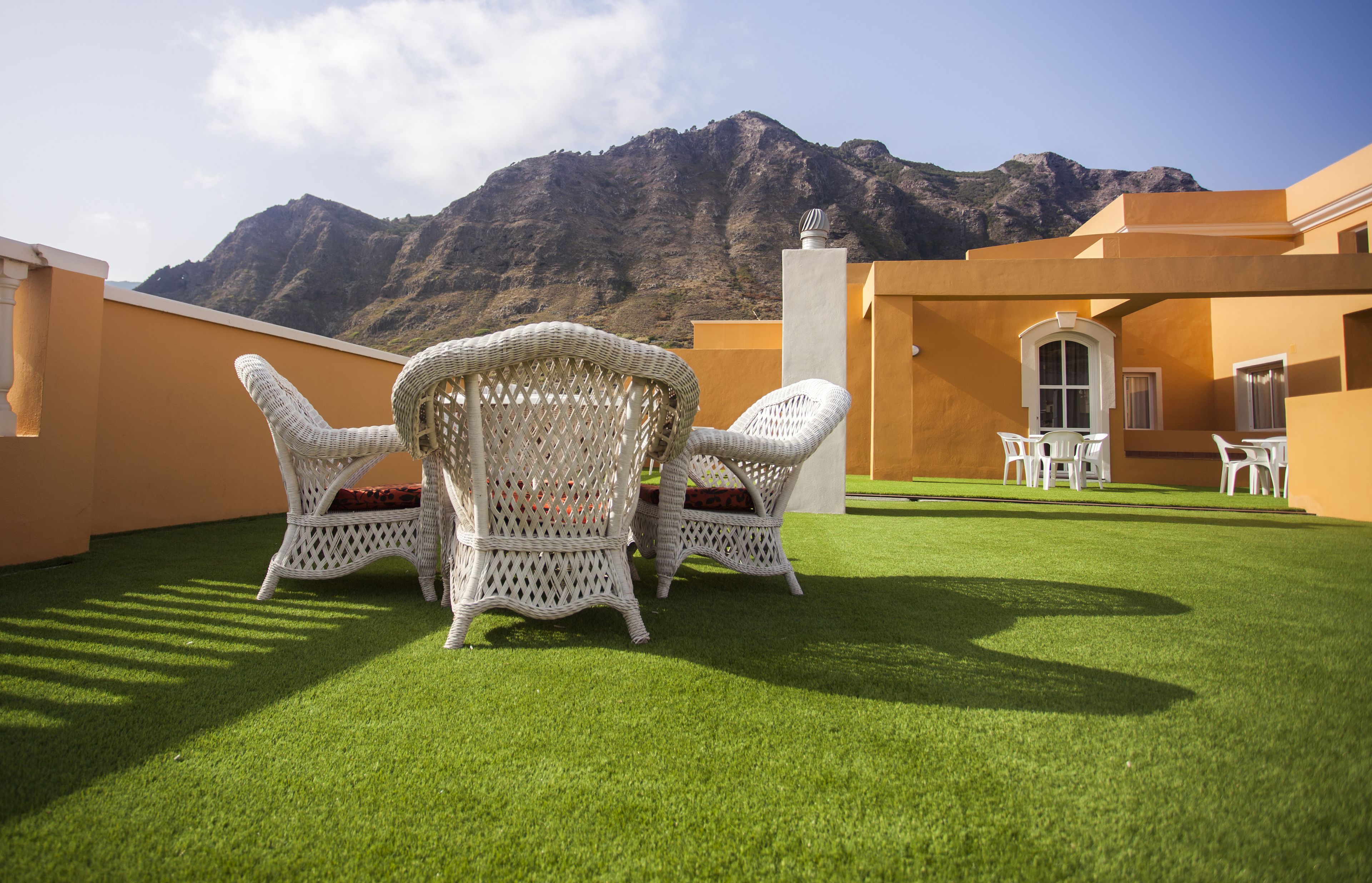 Una terraza con sillas de mimbre y una mesa, un paisaje de montaña al fondo y un edificio con paredes amarillas.