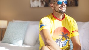 Eine Person sitzt auf einem Bett, trägt ein gelbes T-Shirt und eine Sonnenbrille, neben ihr ein Kopfkissen und ein Bild an der Wand.