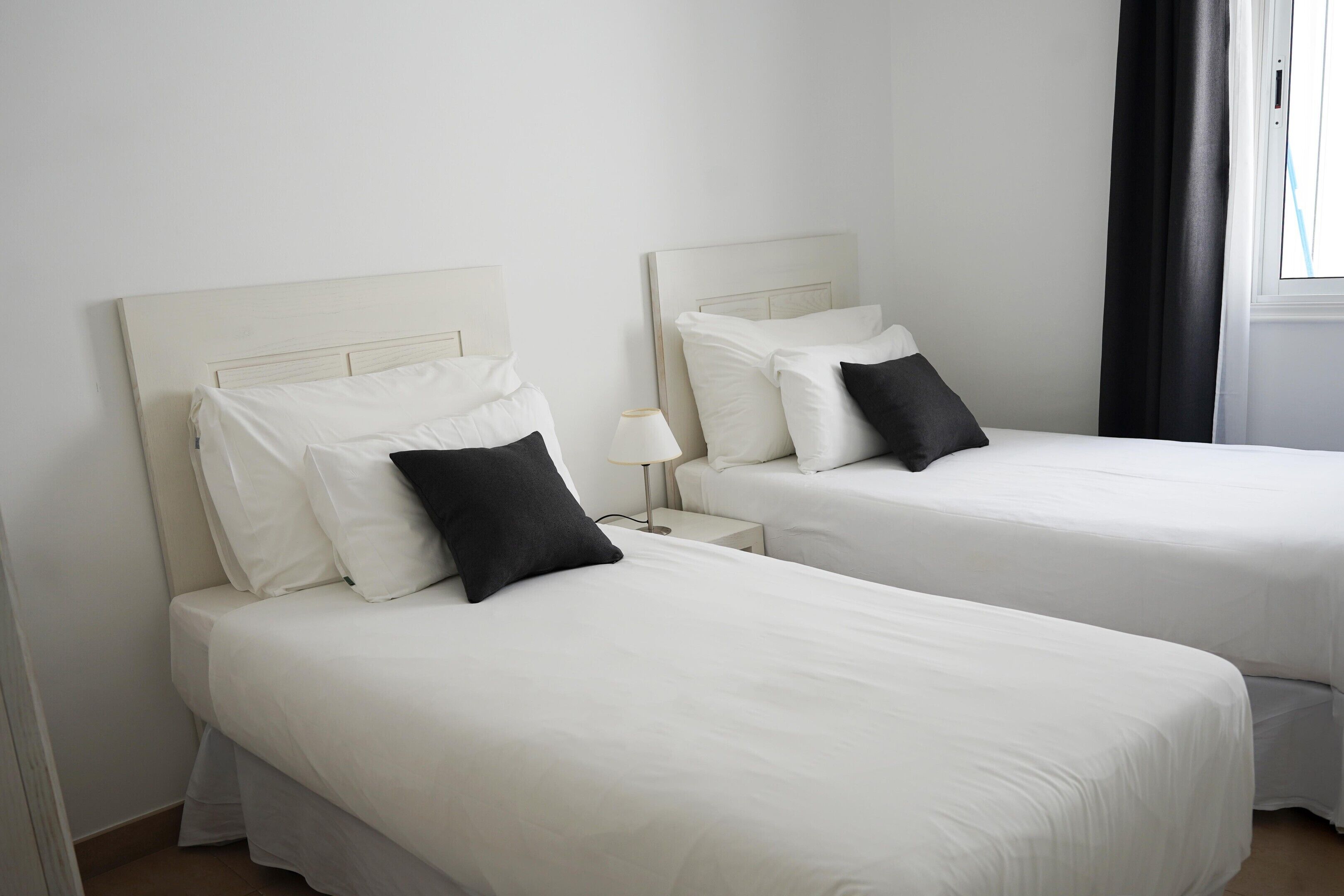 Deux lits simples avec du linge de lit blanc et des oreillers noirs, une table de chevet avec une lampe, et une fenêtre avec un rideau noir.