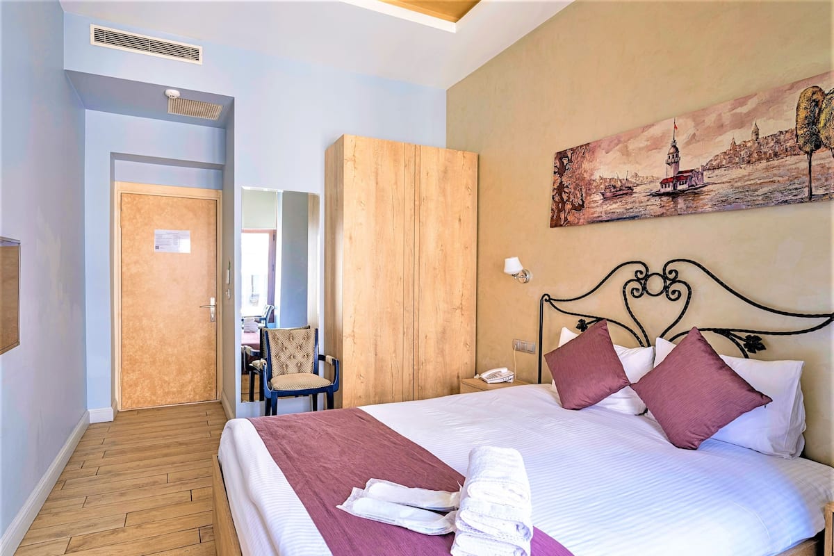 A hotel room with a bed, a wooden wardrobe, a chair, a nightstand with a lamp, and a painting on the wall.