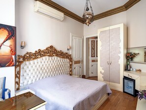 A bedroom with a tufted headboard, a bed with a light-colored bedspread, a small bedside table, a nightstand, a wall-mounted air conditioner, a chandelier, a door with a glass panel, a mirror, and a small cabinet with a mirror and flowers. - Ragıp Pasha Apartments (Istanbul)