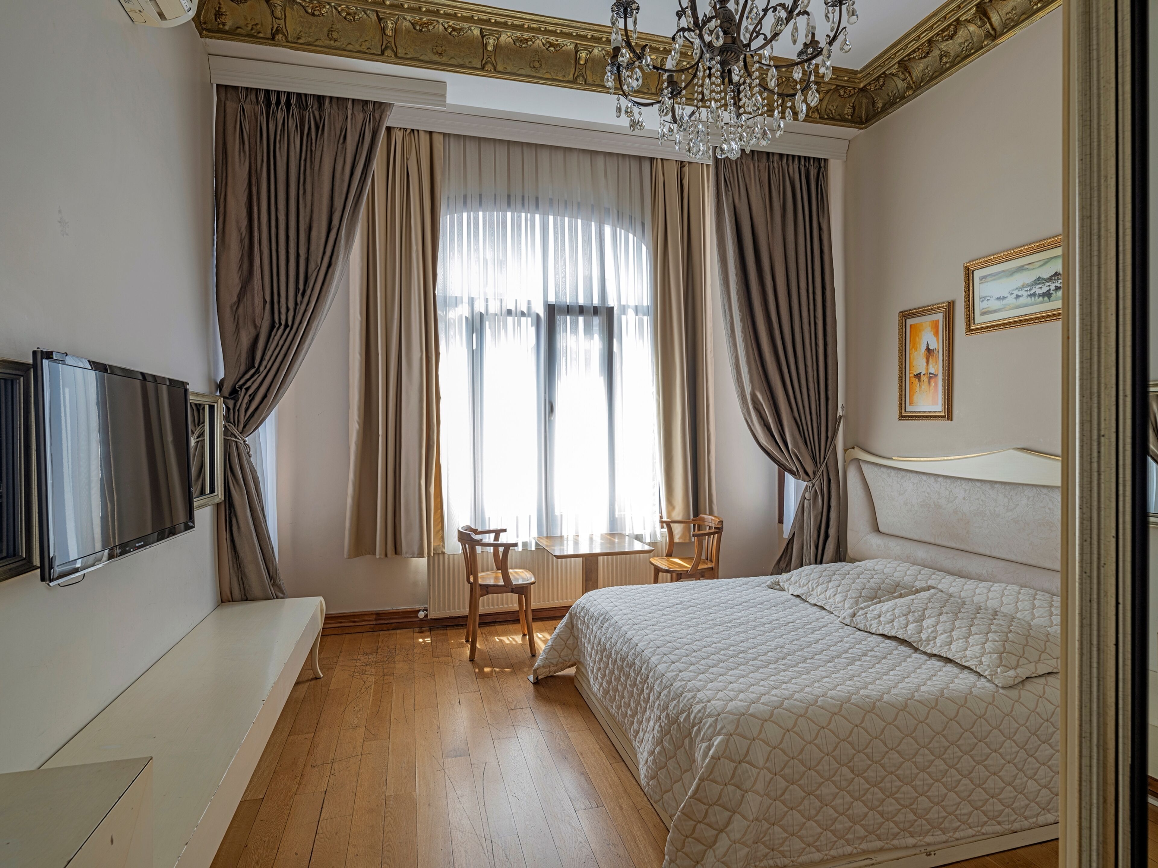 A bedroom with a bed, a desk, and a chair. There is a window with curtains, a chandelier, and a painting on the wall.
